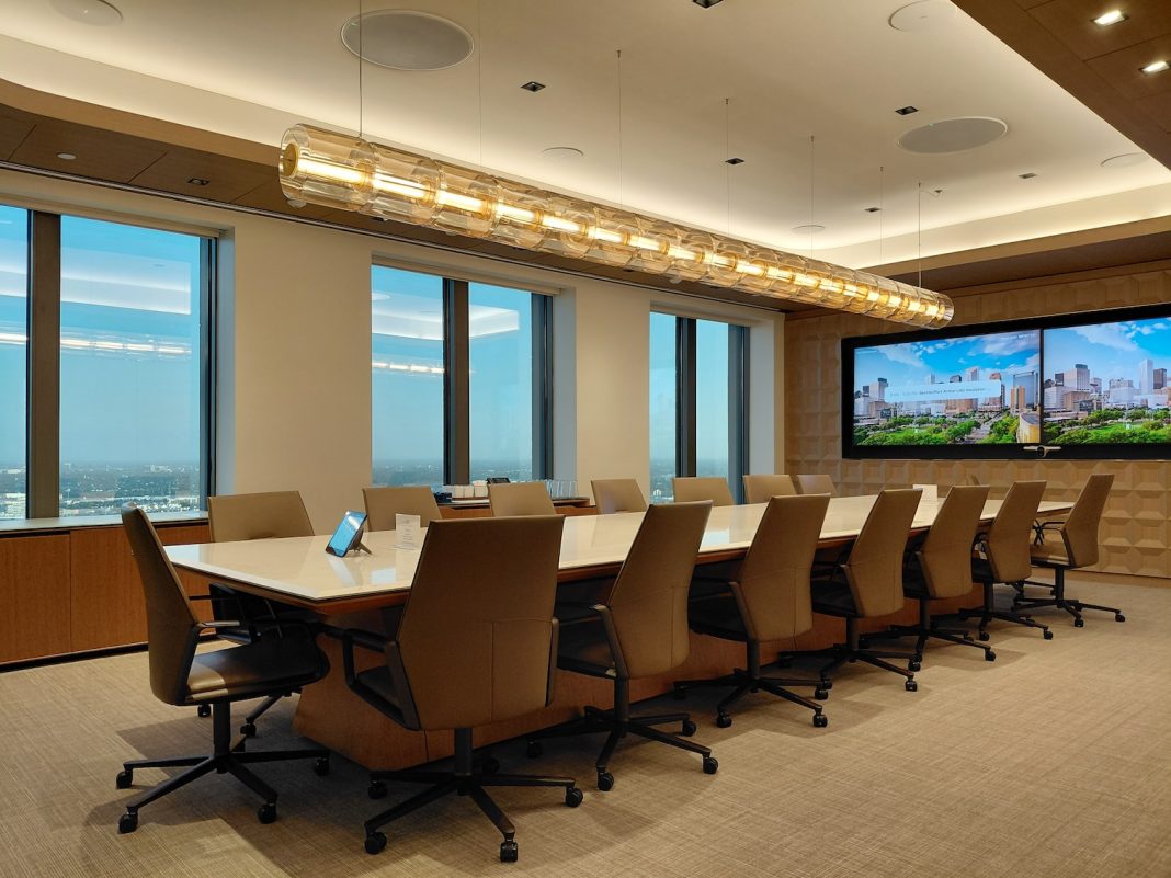 A modern conference room with a long table, leather chairs, large windows overlooking a city, and dual screens displaying a cityscape image.